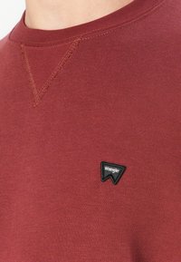 Maroon sweatshirt with a round neck and a textured v-stitch detail. Features a black triangular Wrangler logo patch on the lower front.