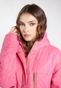 Jachetă roz matlasată cu glugă, având accesorii din metal auriu, două buzunare cu fermoar și o textură netedă. Închidere cu fermoar cu accente din piele.