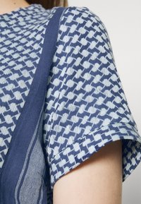 Kortärmad topp med ett strukturerat blått och vitt geometriskt mönster, med en kontrasterande marinblå kant längs axeln och halsringningen.