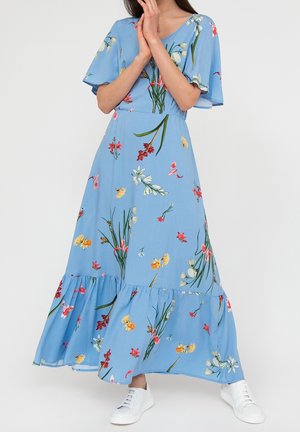 Kobieta ubrana w niebieską sukienkę midi w kwiaty z falbanowymi rękawami i białe trampki, stojąca na jednolitym tle.