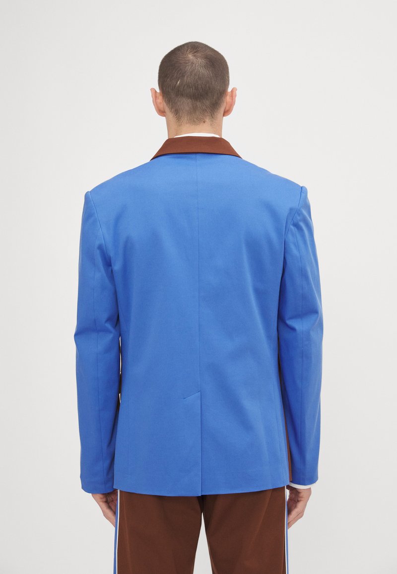 Blazer blu con colletto marrone, realizzato in tessuto liscio. Presenta uno spacco singolo e una vestibilità sartoriale. Indossato sopra pantaloni marroni.