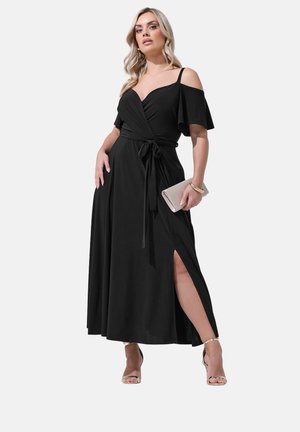 Kvinde iført sort skulderløs omslagskjole med sideslids, holder en beige clutch og bærer en guldbillig og hælede sandaler.