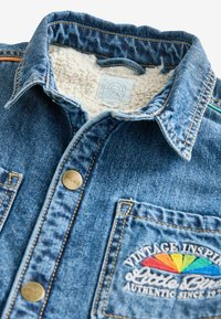 Jeans srajca s bež flisom notranjosti, kovinskimi gumbi, izvezenim logotipom na žepu za prsi in kontrastnimi šivi.