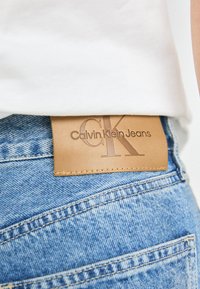 Tan spalvos odos etiketė su išspaustu užrašu "Calvin Klein Jeans" virš mėlynų džinsų, demonstruojančių nusidėvėjusią tekstūrą ir matomus siuvimo detalių.
