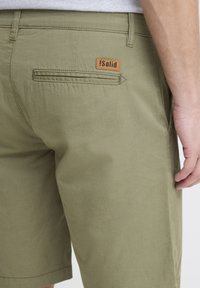 Olivgrüne Shorts mit glatter Textur, ausgestattet mit einer Gesäßtasche und einem braunen Leder-Label. Klassischer Schnitt mit klaren Linien.