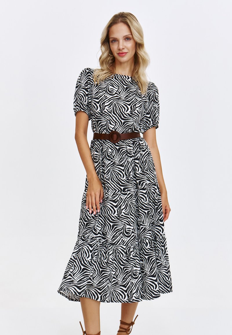 Modelka pozując w sukience w czarno-białe paski zebry z bufkami i brązowym paskiem, prezentuje stylowy, nowoczesny look.