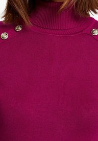 Femme portant un pull à col roulé côtelé magenta avec des boutons décoratifs dorés sur les coutures des épaules.