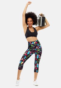 Top corto atlético negro y leggings de cintura alta con un colorido diseño geométrico. La modelo sostiene un altavoz portátil y lleva zapatillas blancas.