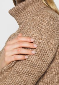 Ljusbrun stickad tröja med hög ribbad krage, med ett texturerat mönster. En hand som vilar på axeln ger extra detalj.