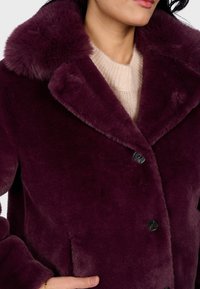 Mujer vestida con un abrigo de piel sintética morado oscuro con un gran cuello sobre un suéter de punto beige, con una mano en el bolsillo del abrigo.