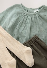 Haut vert clair avec plis et broderies florales, chaussettes côtelées crème, et pantalon en velours côtelé vert olive à taille élastique.