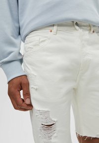 Pantalones cortos de mezclilla blanca con áreas desgastadas y bordes deshilachados. Detalles de herrajes plateados con un sudadera azul claro visible en la parte superior.