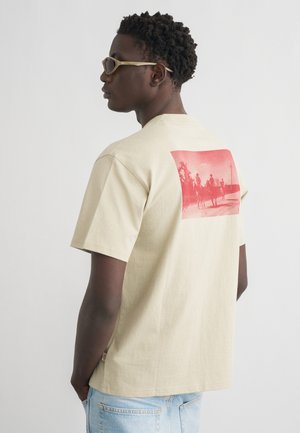 Mann iført beige t-skjorte med rød trykk av tre cowboys på hester, kombinert med lyseblå jeans og beige solbriller, vendt mot venstre.