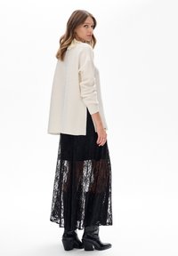 Kobieta ubrana w biały oversize'owy sweter, czarną koronkową spódnicę i czarne botki na obcasie, stoi i spogląda przez ramię na tle białego tła.