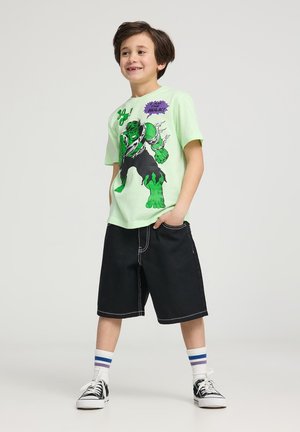 T-shirt verde chiaro con grafica del Hulk in stile fumetto, abbinato a pantaloni corti neri e sneakers. Calzini bianchi a righe blu.