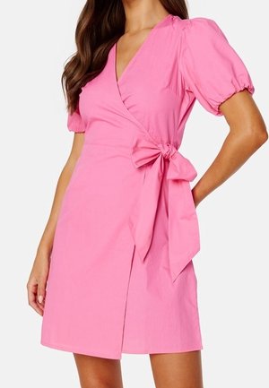 Mujer con un vestido cruzado rosa con mangas abullonadas y un lazo anudado en la cintura, de longitud hasta mitad del muslo, de pie con una mano en la cadera.