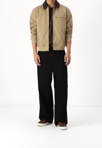 Chaqueta bomber color arena con cuello oscuro y cierre de cremallera, combinada con pantalones de pierna ancha negros y zapatillas marrones con suelas blancas.