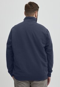 Námornícky modrý sweatshirt s vysokým golierom a rebrovanými manžetami, vyrobený z jemnej látky. Jednoduchý zadný diel so malou nálepkou na krku.