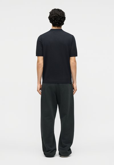 Άνδρας με μπούκλες, σκοτεινά μαλλιά, φοράει μαύρο πόλο με κοντά μανίκια και άνετο σκούρο παντελόνι, στέκεται πλάτη σε απλό φόντο.