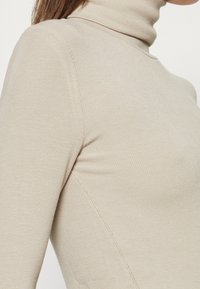 Primo piano di un maglione a collo alto beige a coste indossato da una persona, che mostra texture e dettagli delle cuciture sulla spalla e sul lato.
