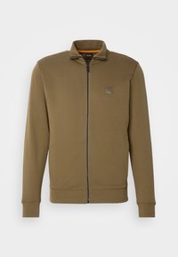 Olivgrön zip-up jacka med hög krage, långa ärmar, ribbade manschetter och liten fyrkantig logotyp på bröstet.