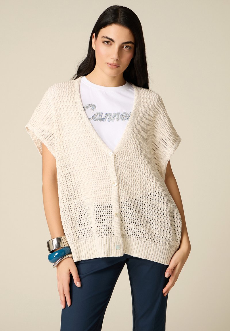 Donna con capelli lunghi e scuri che indossa un gilet lavorato a maglia color crema, morbido e con bottoni, sopra una maglietta bianca e pantaloni blu scuro, accessoriata con braccialetti spessi.