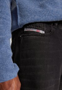Blå strikkegenser med ribbede mansjetter, paret med svarte denimjeans som har en merkeetikett og en teksturert finish. Nærbilde.