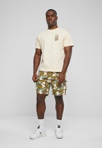Beige t-shirt med et trykt mærke på brystet, parret med grønne camo-shorts med lommer, afsluttet med hvide sneakers og sokker.