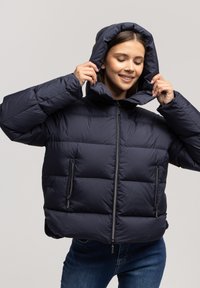 Młoda kobieta uśmiechająca się przy okazji dostosowywania kaptura swojej granatowej kurtki puchowej, nosząca niebieskie dżinsy na jednolitym tle.