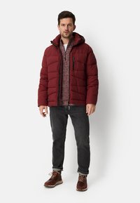 Bordowa pikowana kurtka z kapturem, z kieszenią na piersi na zamek oraz fakturowaną warstwą dzianiny pod spodem, zestawiona z ciemnymi jeansami i brązowymi butami.