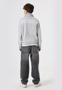 Grijze gebreide trui met hoge kraag met de tekst "ATELIER OFF ROAD" op de rug, gecombineerd met zwarte spijkerbroek en witte sneakers.