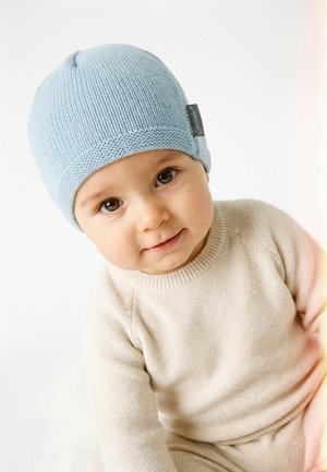 Toddler in beige knit outfit and light blue knit hat, looking toward the camera with a slight smile on a white background.