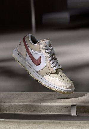 Beżowo-biały sneaker z czerwonym logo Nike i białymi sznurowadłami, przedstawiony w powietrzu nad betonową powierzchnią z delikatnym cieniem w tle.