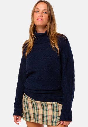 Marineblauer Kabelstrickpullover mit hohem Kragen und geripptem Saum. Verfügt über strukturierte Muster und locker sitzende Ärmel. Kombiniert mit einem karierten Rock.