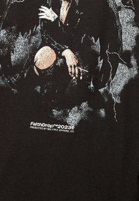 Persona con ropa oscura sentada con los brazos cruzados y una rodilla doblada, sosteniendo un cigarrillo, rodeada de patrones abstractos en negro y gris.
