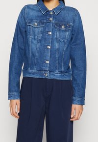 Giubbotto di jeans in blu medio con una vestibilità strutturata e slim, dotato di colletto, chiusura a bottoni, due tasche sul petto e dettagli in metallo argentato.