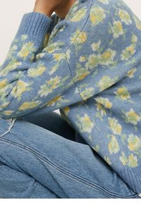 Personne portant un pull bleu avec des motifs floraux jaunes et blancs et un jean bleu clair, assise les bras reposant sur les genoux.