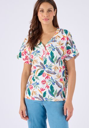Donna che indossa una camicetta bianca a maniche corte con stampa colorata di foglie e fiori tropicali e jeans azzurri.