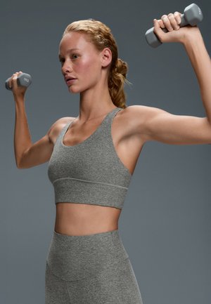 Une femme en tenue de sport grise lève deux haltères et contracte les bras devant un fond gris uni.