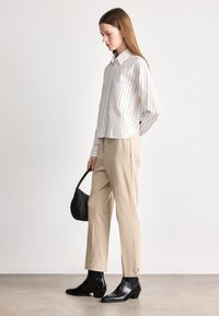 Camisa de botões às riscas brancas e bege, calças bege ajustadas e botas pretas de tornozelo. Pequena mala preta. Design simples com texturas subtis.