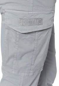 Jasnoszare spodnie cargo z klapką, na której znajduje się logo "Schott NYC" w szarym hafcie. Gładki, trwały materiał z widocznymi szwami.