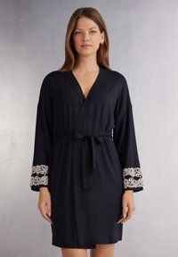 Marineblauwe ochtendjas met een strikceintuur, voorzien van lange mouwen met witte kantaccenten bij de manchetten. Zacht materiaal en een knielange pasvorm.