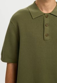 Homme portant un polo en tricot à manches courtes vert olive avec un col et trois boutons marron.
