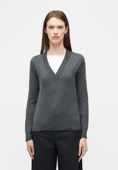 Femme aux cheveux bruns et raides, portant un pull gris à col en V sur un haut blanc et un pantalon noir, debout devant un fond blanc uni.
