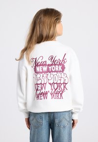 Personne avec de longs cheveux portant un sweat blanc avec le texte "New York" en couches de couleur rose et violette, associé à un jean bleu clair.