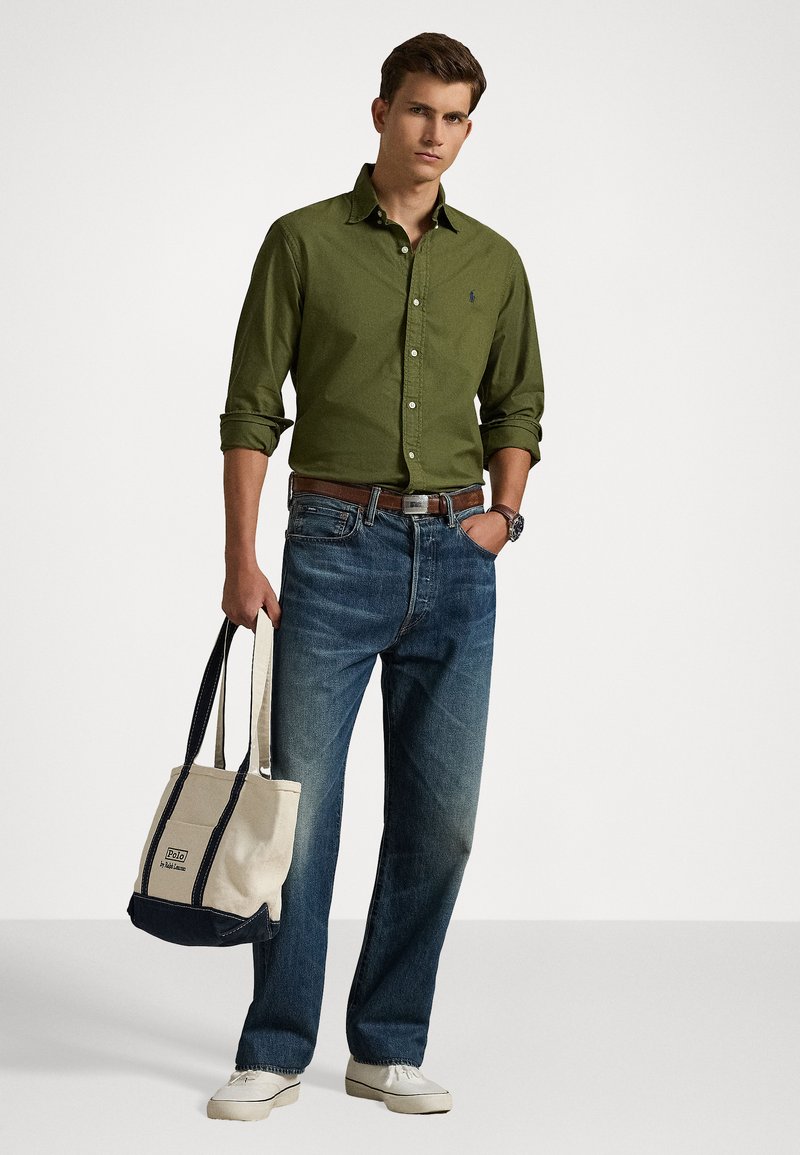 Green button-up shirt, dark jeans, beige tote bag with navy accents, and white shoes. Features a watch and belt. Casual outfit.