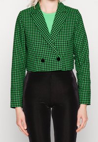 Vihreä ja musta houndstooth-tyylinen lyhyt blazer, jossa on vaaleat kaulukset, kaksinkertaiset napit ja pitkät hihat, yhdistettynä mustiin ribbihousuihin.