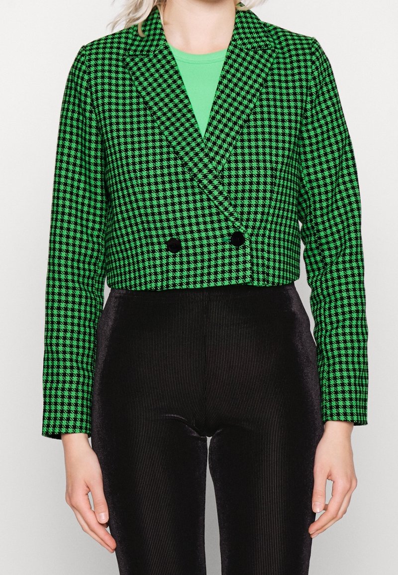 Vihreä ja musta houndstooth-tyylinen lyhyt blazer, jossa on vaaleat kaulukset, kaksinkertaiset napit ja pitkät hihat, yhdistettynä mustiin ribbihousuihin.