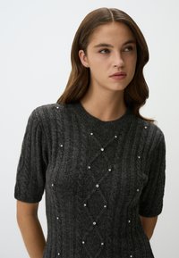 Pull en maille gris foncé à manches courtes présentant un motif texturé avec des formes de diamant et des strass décoratifs éparpillés.
