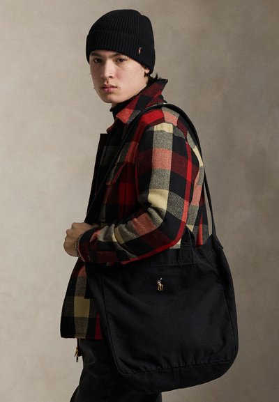 Młoda osoba nosząca czarną czapkę typu beanie oraz czerwoną, czarną i beżową kratowaną kurtkę, niosąca czarną torbę na ramię z małym haftowanym logo.